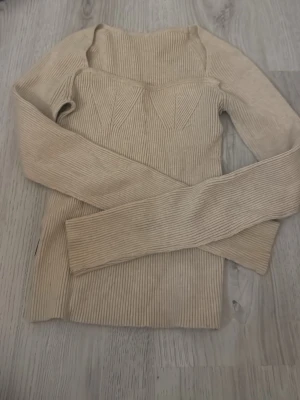 Beige ribbstickad tröja - Trendig beige ribbstickad tröja med lång ärm och markerad byst. Tröjan har en kroppsnära passform och bred, fyrkantig urringning. Perfekt att styla till jeans eller kjol för en snygg vardagslook.