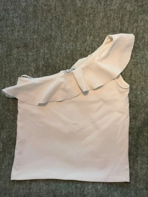 Vit oneshoulder topp med volang - Säljer en vit oneshoulder topp med bred volang längs halsringningen. Nästan aldrig använd, inte kommit till användning 😕37 cm bred, 38 cm lång.