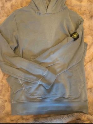Blå hoodie från Stone Island Junior - Snygg blå hoodie från Stone Island Junior med klassisk huva och känguruficka. Ikonisk svart och gul logotyp på vänster ärm. Tillverkad i mjukt bomullsmaterial som känns skönt mot huden. Perfekt för en avslappnad och trendig stil.