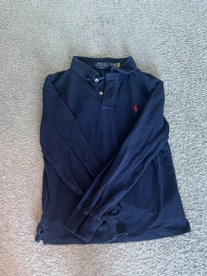 Mörkblå långärmad piké från Polo Ralph Lauren - Snygg mörkblå långärmad pikétröja från Polo Ralph Lauren i slim fit. Klassisk krage, knappar framtill och ikonisk röd logga broderad på bröstet. Perfekt för en stilren och avslappnad look. Detta är en barnstorlek men det är den största barnstorleken L(14-16) G! Vilket betyder att den passar xs som vanlig storlek och möjligtvis small