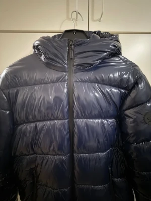 Carrera Jeans mörkblå glansig dunjacka  - Snygg, varm och glansig mörkblå dunjacka från Carrera Jeans! Överlag i väldigt fint skick med smidiga bärremmar på insidan.      Obs: Liten reva i ena innermudden, men den döljs helt under ärmen när jackan bärs (se bilder)  