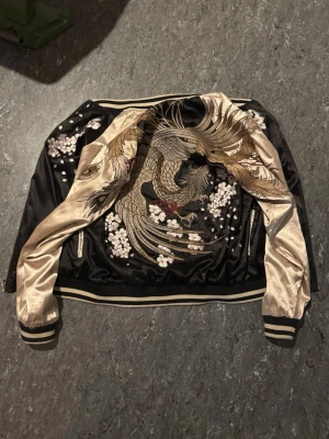 Svart och guld bomberjacka med brodyr från Kidoriman - Snygg svart och guld bomberjacka i satin med broderade körsbärsblommor och en stor drake på ryggen. Den är från Kidoriman och säljs inte längre där. Är i princip oanvänd och det är äkta brodering. Det står storlek ”M” men jag är storlek S/XS och den passar mig. Köpte den för cirka 1300kr
