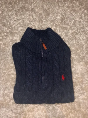 Mörkblå kabelstickad tröja med halvzip - Mörkblå kabelstickad tröja från Polo Ralph Lauren med halvzip och hög krage. Tröjan har ett rött broderat logotyp på bröstet och ribbade muddar. Perfekt för lager på lager och riktigt snygg till jeans.