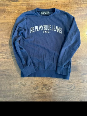 Mörkblå sweatshirt från Replay - Mörkblå sweatshirt från Replay med vit text 'REPLAY BLUE JEANS INC.' på bröstet. Klassisk rund halsringning och ribbade muddar vid ärmslut och nederkant. Perfekt till jeans för en avslappnad stil.