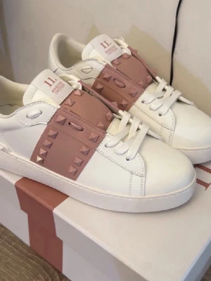 Vita sneakers med rosa nitar - Snygga vita sneakers med bred rosa rem över vristen dekorerad med matchande nitar. Skorna har vita snören, platt sula och detaljer i både vitt och rosa. Perfekta för dig som vill ha en unik och trendig look.