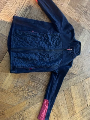 Mörk blå ridflis från Decathlon med quiltad front - Snygg mörkblå jacka från Decathlon med quiltad frontpanel och dragkedja. Jackan har långa ärmar med en rosa detalj på ena ärmen och praktiska fickor med dragkedja. Perfekt för kyliga dagar och sportiga aktiviteter.