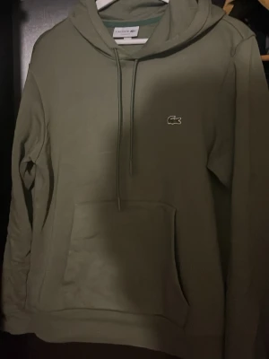 Olivgrön hoodie från Lacoste - Snygg olivgrön hoodie från Lacoste med klassisk krokodillogga på bröstet. Hoodien har huva med dragsko, stor magficka och ribbade muddar. Perfekt för en chill och avslappnad stil.