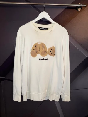 Palm angels tröja  - Vit stickad tröja från Palm Angels med ikoniskt nallebjörnsmotiv på bröstet och svart logotyptext under. Tröjan har rund halsringning och långa ärmar. Perfekt för dig som gillar streetwear och vill ha något unikt i garderoben.