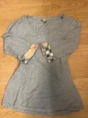 Grå långärmad topp Burberry Brit - Snygg grå långärmad topp från Burberry Brit med klassiska rutiga detaljer på ärmsluten. Tillverkad i mjuk bomull och har en avslappnad passform. Perfekt för dig som gillar stilrena plagg med en twist.