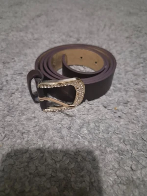 Brunt bälte med glittrande spänne - Snyggt brunt bälte i läderimitation med ett silvrigt spänne dekorerat med små glittrande stenar. Perfekt accessoar för att lyfta din outfit med lite extra bling. Smalt och stilrent, passar till både jeans och kjol.