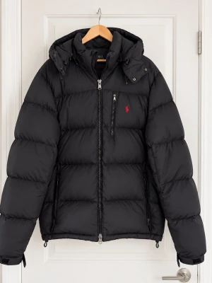 Svart dunjacka från Polo Ralph Lauren - Snygg svart dunjacka från Polo Ralph Lauren med röd broderad logga på bröstet. Jackan har huva, dragkedja framtill och två sidofickor med dragkedja. Perfekt för kalla vinterdagar och riktigt stilren look.