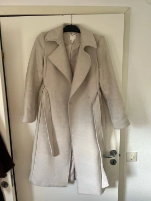 Beige lång kappa med skärp - Stilren beige kappa med bred krage och knytskärp i midjan. Kappan har långa ärmar, två sidofickor och en rak passform. Perfekt för kyligare dagar och ger en klassisk look till din outfit.