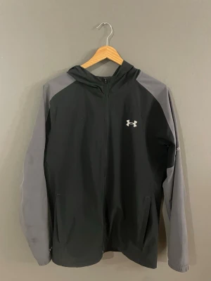 Under Armour svart och grå vindjacka - Snygg vindjacka från Under Armour i svart med grå ärmar och huva. Jackan har dragkedja framtill, två sidofickor och en vit logga på bröstet. Perfekt för blåsiga dagar och sportiga outfits. Använd fåtal gånger. Säljer eftersom att den inte kommer till användning längre 