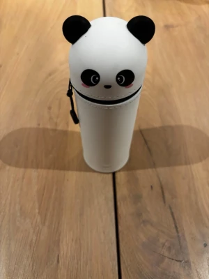 Pandaformad pennfodral med dragkedja - Ett vitt cylinderformat pennfodral med svart dragkedja och lock utformat som ett pandaansikte med öron. Fodralet har ett litet handtag på sidan och är gjort i plast eller silikon. Perfekt för att förvara pennor eller småprylar.