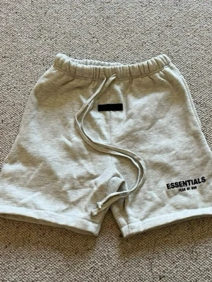 Essentials shorts  - Ett par gråa essentials shorts med svart text nere vid vänstra benet. De är i storlek M och i nytt skick. Pris kan diskuteras och skriv om du har några frågor. 