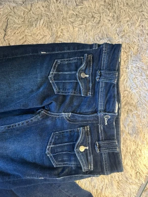 Mörkblå jeans med fickdetaljer - Säljer ett par mörkblå jeans med unika fickor framtill som har lock och knapp. Jeansen har breda hällor och klassiska bakfickor utan lock. Snygga kontrastsömmar och rak passform. Perfekta för dig som vill ha något lite annorlunda!