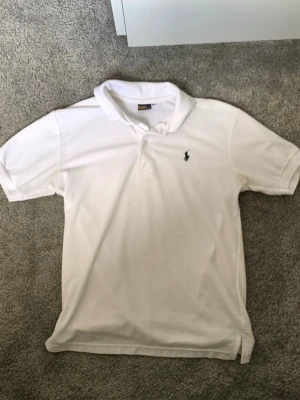 Vit pikétröja från Ralph Lauren - Klassisk vit pikétröja från Ralph Lauren med blå logga broderad på bröstet. Tröjan har korta ärmar, krage och knäppning framtill. Tillverkad i mjukt bomullsmaterial som är skönt mot huden. Perfekt till jeans eller shorts för en clean look.