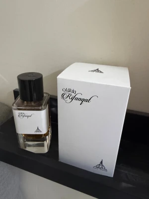 Rafaqat parfym med elegant flaska - Upptäck Rafaqat parfym i en stilren, fyrkantig glasflaska med svart lock och vit etikett. Doften kommer i en matchande vit kartong med svart text och dekorativ logga. Perfekt för dig som gillar exklusiva och unika parfymer.