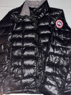 Svart jacka från Canada Goose - Snygg svart regnjacka från Canada Goose med quiltad design och hög krage. Jackan har ett klassiskt märke på bröstet och dragkedja framtill. Materialet är glansigt och vattenavvisande, perfekt för regniga dagar.