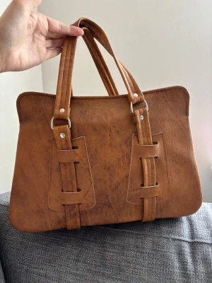 Brun fyrkantig handväska i konstläder - Snygg brun handväska i konstläder med rektangulär form och dubbla handtag. Väskan har dekorativa sömmar och metallnitar, samt en unik design med breda remmar framtill. Perfekt för dig som gillar stilrena accessoarer med en twist. Mått 35x25x14 cm 
