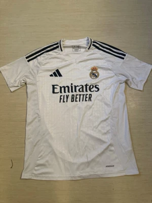 Real Madrid matchtröja Mbappé 9 - Vit Real Madrid matchtröja med svarta detaljer och klubbmärke på bröstet. Adidas-logga och sponsortryck på framsidan, samt Mbappé och nummer 9 på ryggen. Tillverkad i lätt och ventilerande material, perfekt för fotbollsträning eller match.