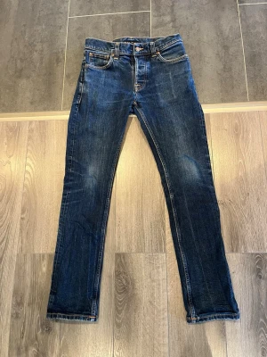 Nudie Jeans Grim Tim 31/32 - Riktigt snygga Nudie Jeans Grim Tim i mörkblå denim med naturliga fades och grym patina. Klassisk slim/straight passform som sitter väldigt bra. Orange kontrastsömmar och den ikoniska Nudie-detaljeringen baktill.  Jeansen är använda men i fint skick och har fått en unik vintage-look med tiden. Perfekta om du gillar raw denim, archive/grisch-stil eller jeans med karaktär.  Modell: Grim Tim Material: 99% bomull + elastan Mått kan skickas vid intresse 📏 Snabb affär prioriteras!