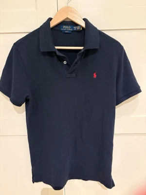 Marinblå piké från Polo Ralph Lauren - Klassisk marinblå pikétröja från Polo Ralph Lauren . Tröjan har korta ärmar, krage och två knappar framtill samt loggan på bröstet. Använd fåtal gånger och är som ny. Köpt på kidsbrandstore. Storlek XL i juniorstorlek