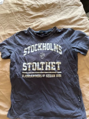 Blå Djurgården t-shirt Stockholms Stolthet - Säljer en blå t-shirt med texten 'Stockholms Stolthet' och Djurgårdens IF-emblem på bröstet. Klassisk rund hals och korta ärmar. Perfekt för dig som vill visa ditt stöd för Djurgården på ett stilrent sätt.
