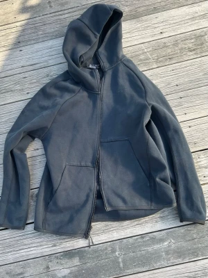 Mörkgrå zip hoodie från Nike - Snygg mörkgrå hoodie från Nike med dragkedja framtill och två stora fickor. Klassisk huva och diskret Nike-logga på bröstet. Tillverkad i mjukt bomullsmaterial som är skönt att ha på sig. Perfekt för en avslappnad streetwear-look.