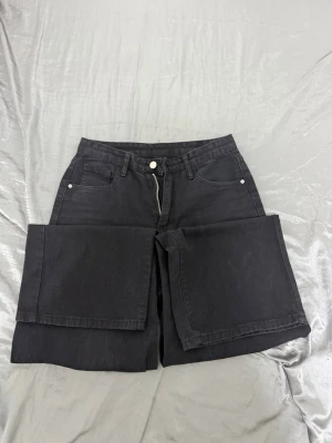 Svarta baggy jeans för tjejer - Säljer ett par svarta denimshorts med klassisk femficksdesign och knappgylf. Shortsen har raka, breda ben och bälteshällor i midjan. Perfekta för dig som gillar en avslappnad och stilren look.