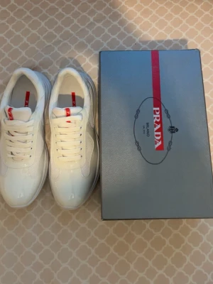 Vita sneakers från Prada - Stilrena vita sneakers från Prada med röd logga på plösen. Skorna har en chunky sula, meshpaneler och detaljer i blankt material. Klassisk snörning och modern design som passar till många outfits.