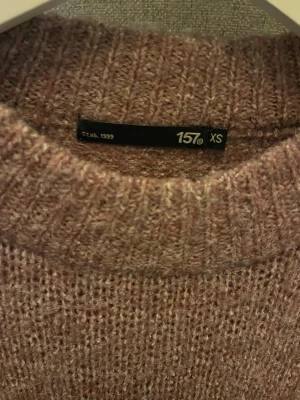 Rosa stickad tröja från 157 - Mysig stickad tröja i ljusrosa från 157 med rund halsringning och ribbade muddar. Tröjan har en avslappnad passform och är perfekt för kalla dagar. Materialet känns mjukt och behagligt mot huden.