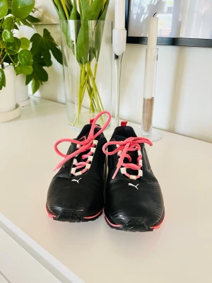 Golfsko Puma svarta sneakers med rosa snören - Svarta sneakers från Puma med vita detaljer och rosa snören. Skorna har låg profil, klassisk logga på tån och är tillverkade i syntetmaterial. Perfekta för dig som gillar sportig och stilren design.