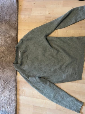 Grå stickad tröja med rund hals - Mysig grön stickad tröja med rund halsringning och ribbade muddar vid ärmslut och nederkant. Tröjan har en enkel och stilren design, perfekt för lager på lager och passar till det mesta. Materialet ser ut att vara mjukt och värmande. Noppar av innan ja skickar! Tar alla bud!