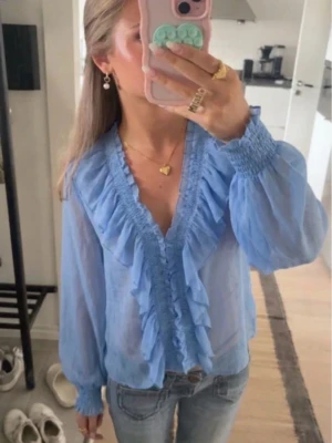 Ljusblå volangblus med v-ringning - Snygg ljusblå blus med v-ringning och volangdetaljer framtill. Blusen har långa, transparenta ärmar med smock vid ärmslut och en luftig, feminin passform. Perfekt att styla med jeans eller kjol för en trendig look.