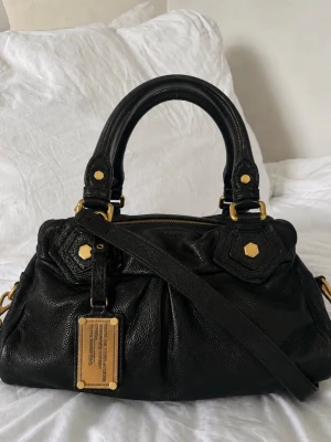 Svart handväska Marc by Marc Jacobs - Snygg svart handväska i läder från Marc by Marc Jacobs med guldfärgade metalldetaljer och dubbla handtag. Väskan har avtagbar axelrem och dragkedja upptill. Insidan är fodrad med ett coolt mönster i svart och vitt med logotyptryck.