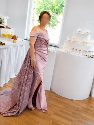 Ljusrosa långklänning med slits - Elegant ljusrosa långklänning med offshoulder-design och slits framtill. Klänningen har en glittrig, broderad detalj och ett långt släp som ger extra wow-faktor. Perfekt för bal eller festliga tillfällen.