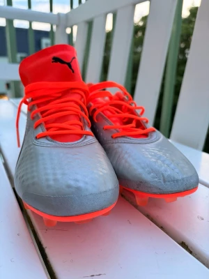 Puma silverröda fotbollsskor stl 40 - Snygga fotbollsskor från Puma i silver med röda detaljer, snörning och dobbar. Ovandelen är i syntetmaterial och skon har en högre plös med Puma-logga. Perfekta för dig som vill sticka ut på planen.