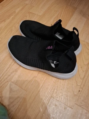 Svarta slip-on sneakers från Fila - Svarta stickade slip-on sneakers från Fila med vit sula och lila logga på plösen. Skorna har en sportig och modern design med elastisk ovandel i textil som ger en skön passform. Perfekta för en avslappnad och trendig stil.