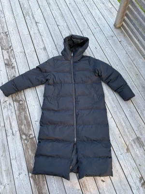 Lång svart dunjacka med huva - Säljer en lång, svart dunjacka med huva och dragkedja hela vägen fram. Jackan har en rak passform, quiltade paneler och två sidofickor. Perfekt för kalla vinterdagar när du vill hålla dig varm och stilsäker.