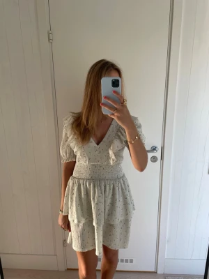 Ljusgrön kortklänning med volang och v-ringning - Söt och somrig ljusgrön volangklänning med små blommiga detaljer, v-ringad hals och volang vid axlarna. Klänningen har korta puffärmar, smock i midjan och dubbla volanglager i kjolen. Perfekt för midsommar, skolavslutning eller svensk sommar.