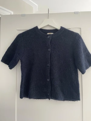 Mörkblå stickad kortärmad kofta - Kortärmad stickad kofta i mörkblått med rund halsringning och knappar framtill. Koftan har en rak passform och är gjord i ett mjukt, lite luddigt material. Perfekt att slänga över en topp för en chill look.