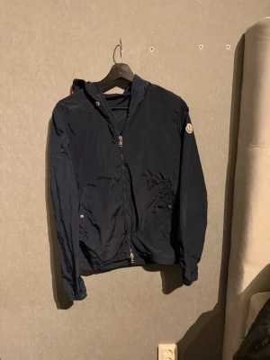 Mörkblå vindjacka med huva - Snygg mörkblå vindjacka med dragkedja framtill och två fickor med tryckknapp. Jackan har huva med röd, vit och blå detalj samt resår vid ärmslut och nederkant. Perfekt för blåsiga dagar och enkel att matcha till olika outfits. (Snöret är borttappat men lär hitta det förhoppningsvis)