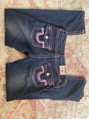 True religion jeans med rosa sömmar - Säljer ett par mörkblå true religion jeans med tydliga rosa kontrastsömmar och dekorativa bakfickor med lock och knapp. Jeansen har en klassisk femficksmodell och raka ben. Det står ingen storlek men har andra i storlek W27 och båda passar ungefär lika. Skriv gärna vid frågor! 