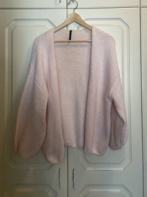 Ljusrosa fluffig kofta - Mysig ljusrosa kofta med öppen front och ballongärmar. Koftan är stickad i ett fluffigt material som ger en mjuk och avslappnad look. Perfekt att slänga över en topp för extra värme och stil.