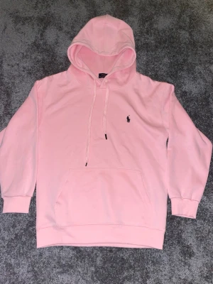 Ljusrosa hoodie med broderad logga - Säljer en ljusrosa hoodie med huva och dragsnören samt en broderad logga på bröstet. Hoodien har en stor magficka och ribbade muddar vid ärmslut och nederkant. Perfekt för en chill och avslappnad stil.