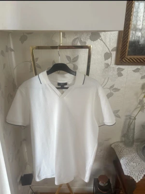 Vit kortärmad henley-skjorta - Snygg vit henley-skjorta med korta ärmar och diskret mörk kant på kragen. Skjortan har en luftig struktur och v-ringad hals. Perfekt för en clean och fräsch stil. 