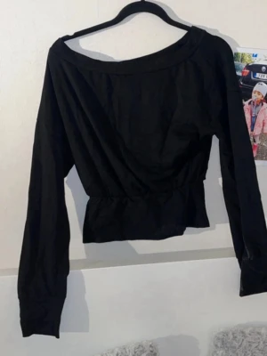 Svart långärmad topp med bred hals - Snygg svart topp med bred, något asymmetrisk halsringning och långa ärmar. Modellen är croppad och har en markerad midja som ger en cool siluett. Perfekt att styla med höga jeans eller kjol för en trendig look.