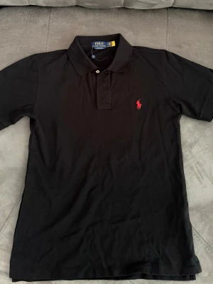 Ralph Lauren - Klassisk svart pikétröja från Polo Ralph Lauren med röd broderad logga på bröstet. Krage och knappar framtill. Perfekt till både jeans och shorts för en clean och stilren look.