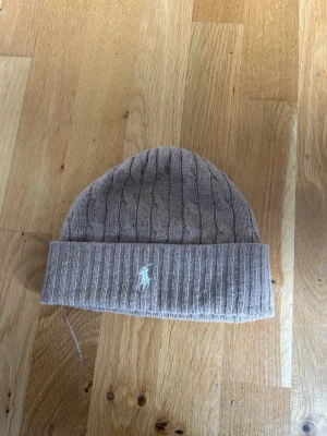 Beige kabelstickad mössa Polo Ralph Lauren - Säljer en beige kabelstickad mössa från Polo Ralph Lauren med vikt kant och den klassiska broderade loggan framtill. Mössan är mjuk och har en stilren design som passar perfekt till höst och vinter. Materialet känns lyxigt och håller värmen bra.